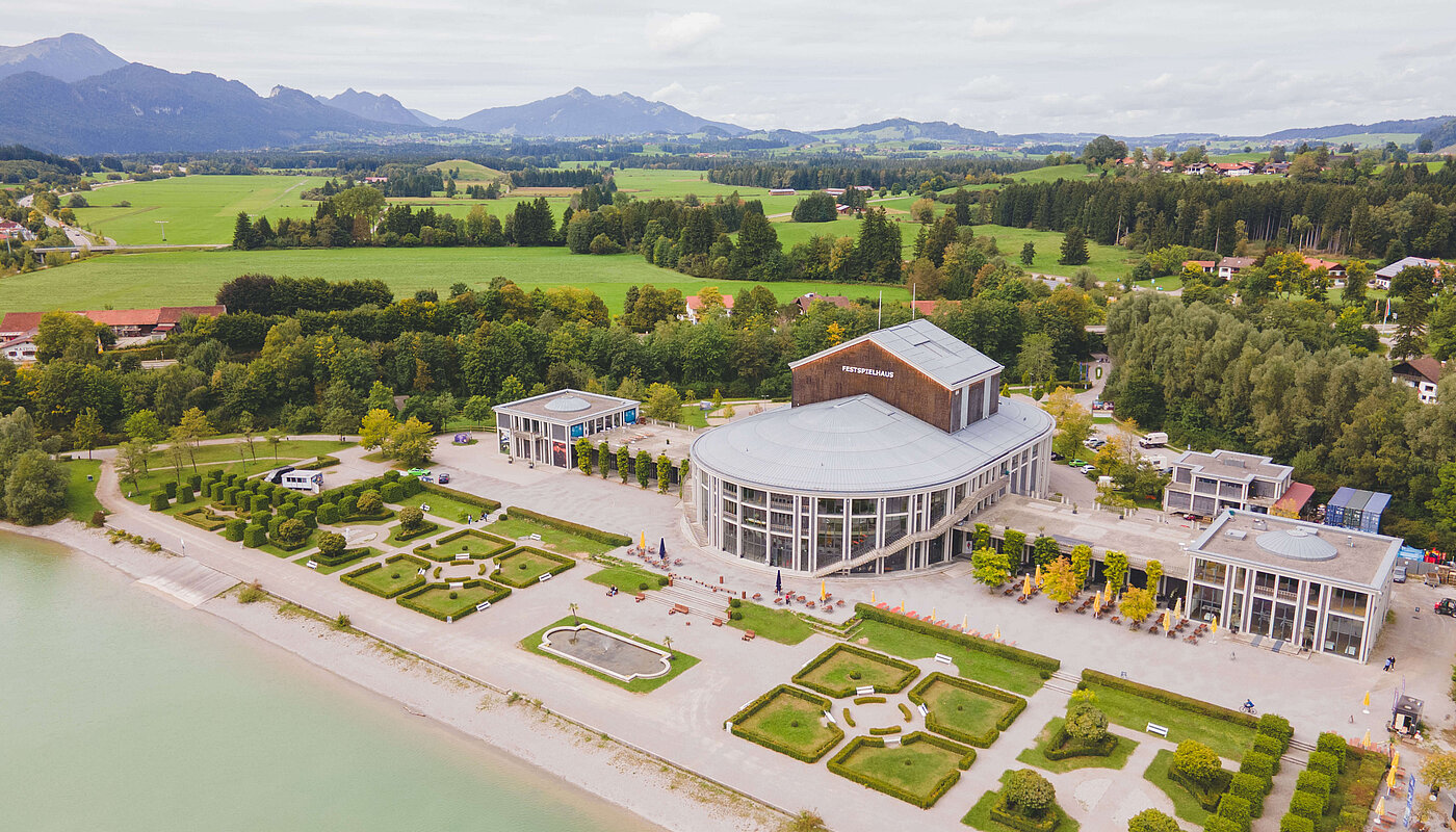 Luftaufnahme eines Gebäudes in der Nähe eines Gewässers, umgeben von Landschaft und Bergen. Im Vordergrund sind gepflegte Gartenanlagen sichtbar. Das Gebäude hat große Fenster und eine moderne Architektur. Der Himmel ist bewölkt, und die Umgebung wirkt grün und natürlich.