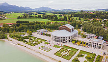 Luftaufnahme eines Gebäudes in der Nähe eines Gewässers, umgeben von Landschaft und Bergen. Im Vordergrund sind gepflegte Gartenanlagen sichtbar. Das Gebäude hat große Fenster und eine moderne Architektur. Der Himmel ist bewölkt, und die Umgebung wirkt grün und natürlich.