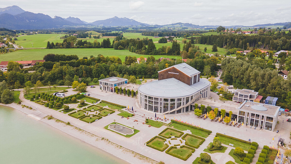 Luftaufnahme eines Gebäudes in der Nähe eines Gewässers, umgeben von Landschaft und Bergen. Im Vordergrund sind gepflegte Gartenanlagen sichtbar. Das Gebäude hat große Fenster und eine moderne Architektur. Der Himmel ist bewölkt, und die Umgebung wirkt grün und natürlich.