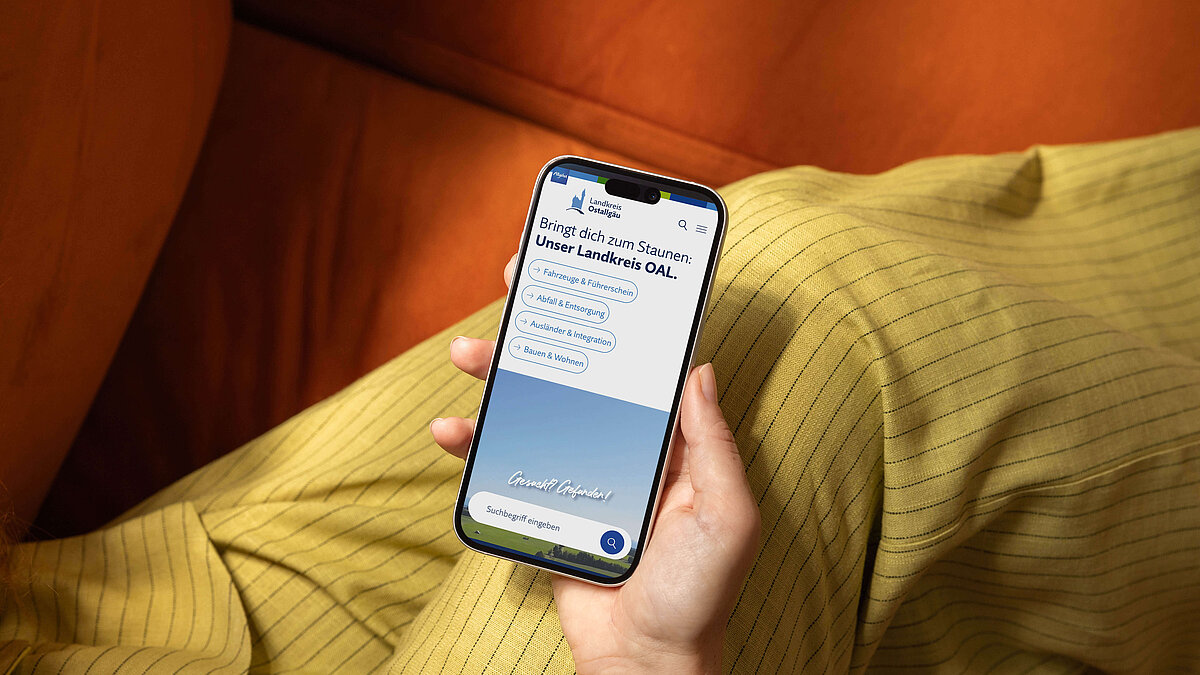 Eine Hand hält ein Smartphone, das eine App mit dem Titel "Bringt die zum Summen: Unser Lernen OAL" zeigt. Der Hintergrund ist eine orangefarbene Couch, und im Vordergrund ist ein gelb gestreifter Stoff sichtbar.