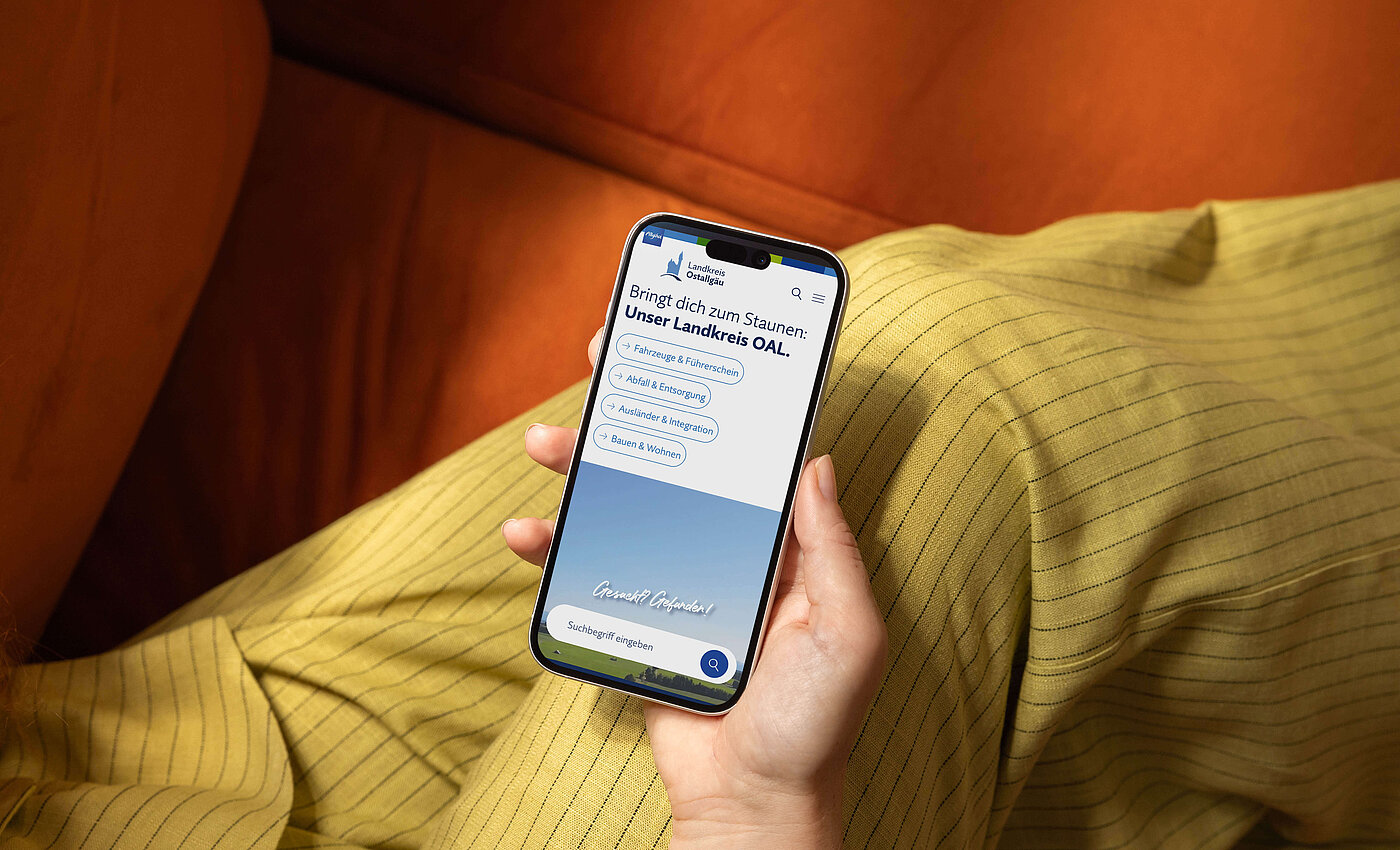 Eine Hand hält ein Smartphone, das eine App mit dem Titel "Bringt die zum Summen: Unser Lernen OAL" zeigt. Der Hintergrund ist eine orangefarbene Couch, und im Vordergrund ist ein gelb gestreifter Stoff sichtbar.