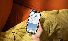 Eine Hand hält ein Smartphone, das eine App mit dem Titel "Bringt die zum Summen: Unser Lernen OAL" zeigt. Der Hintergrund ist eine orangefarbene Couch, und im Vordergrund ist ein gelb gestreifter Stoff sichtbar.