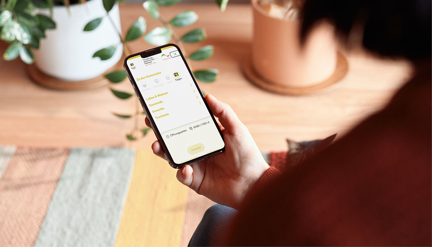 Eine Person hält ein Handy in der Hand, auf dem eine App-Oberfläche zu sehen ist. Im Hintergrund sind Pflanzen und ein farbiger Teppich sichtbar. Die Person hat einen dunklen Pullover an und sitzt auf dem Boden.