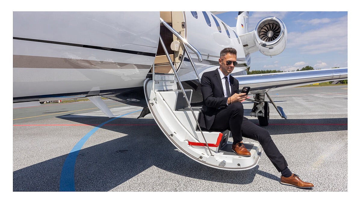 Geschäftsperson sitzt auf der Einstiegstreppe eines Privatjets und checkt E-Mails am Smartphone