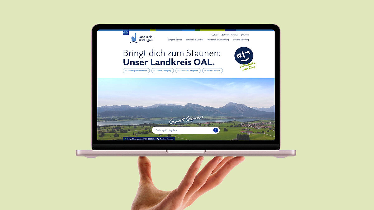 Ein Laptop wird von einer Hand gehalten, auf dem Bildschirm ist eine Webseite zu sehen, die den Landkreis OAL vorstellt. Darauf steht: "Bringt dich zum Staunen: Unser Landkreis OAL" sowie eine Landschaft mit Bergen im Hintergrund.
