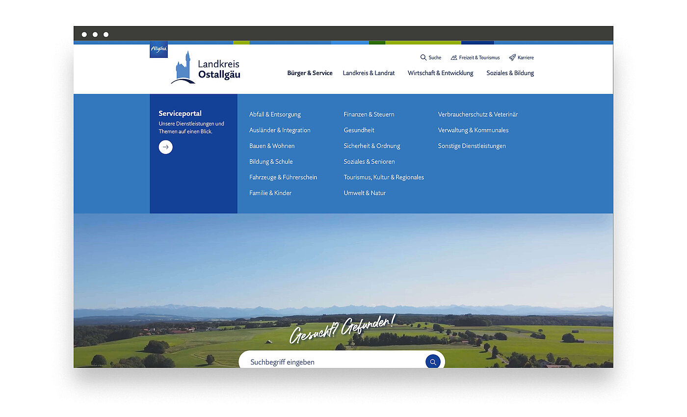 Webseite des Landkreises Ostalgau mit einem blauen Menü oben. Das Hauptthema ist das Serviceportal mit verschiedenen Kategorien wie Familie, Gesundheit und Bildung. Im Hintergrund ist eine Landschaft mit sanften Hügeln und einem klaren Himmel zu sehen.