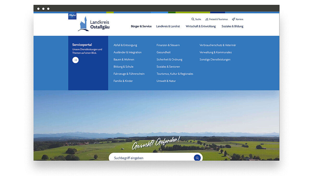 Webseite des Landkreises Ostalgau mit einem blauen Menü oben. Das Hauptthema ist das Serviceportal mit verschiedenen Kategorien wie Familie, Gesundheit und Bildung. Im Hintergrund ist eine Landschaft mit sanften Hügeln und einem klaren Himmel zu sehen.