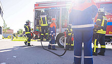 Alt-Text: Eine Gruppe von Feuerwehrleuten arbeitet an einem Feuerwehrfahrzeug. Einige Mitglieder entladen Ausrüstung, während andere mit einem Schlauch beschäftigt sind. Die Szene findet bei Tageslicht statt.