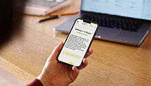 Eine Person hält ein Smartphone in der Hand, auf dem eine Webseite mit dem Titel „Wohnen und Bauen“ angezeigt wird. Im Hintergrund ist ein Laptop und Büromaterial auf einem Tisch sichtbar.