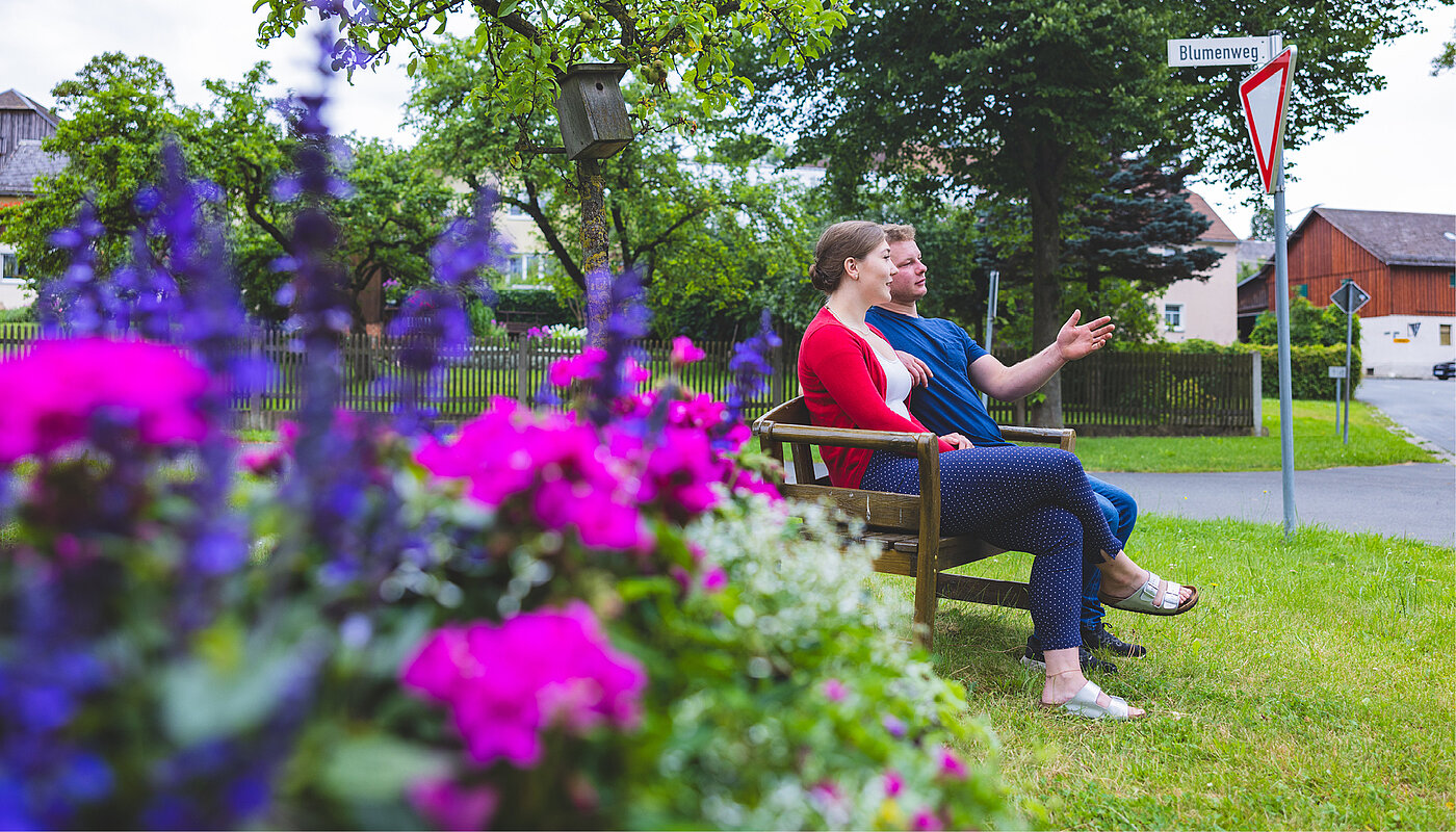 Zwei Personen sitzen auf einer Bank in einem Garten, umgeben von bunten Blumen. Sie blicken entspannt in die Ferne, während im Hintergrund ein Straßenschild zu sehen ist. Die Atmosphäre ist ruhig und einladend.