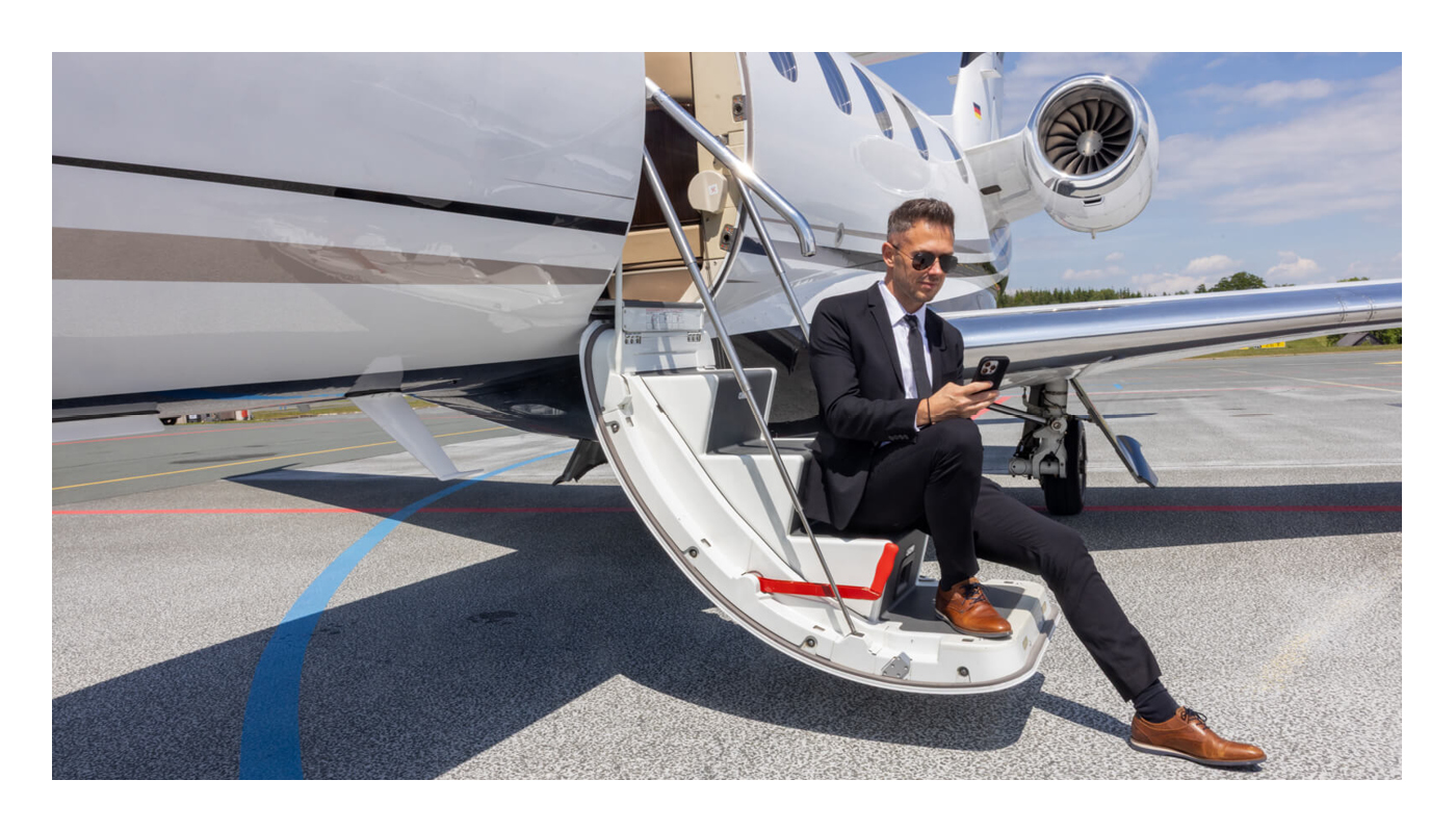 Geschäftsperson sitzt auf der Einstiegstreppe eines Privatjets und checkt E-Mails am Smartphone